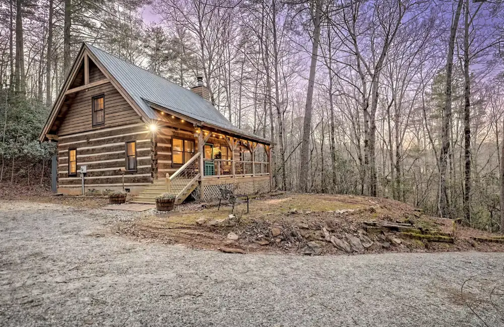 Scott's log cabin, Summer's End, at dusk: warm porch lights glowing on the wraparound porch, with a metal roof and bare hardwood forest under a lavender sky